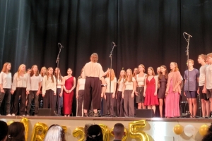 Ein Chor aus vielen Schüler*innen steht auf der Bühne. Eine Chorleiterin steht davor und dirigiert. Die Jugendlichen tragen festliche Kleidung.