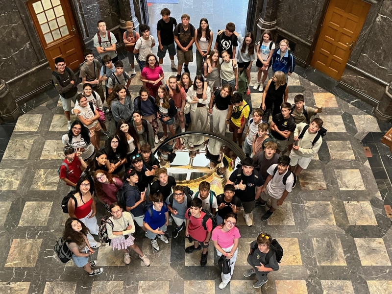 Eine große Gruppe von Jugendlichen steht im Kreis in einem historischen Gebäude. Das Foto wurde von oben aufgenommen. In der Mitte des Raumes liegt eine moderne, runde Glasplatte mit einem Ausstellungsstück darunter. Der Boden ist aus großen, bunten Steinplatten. Um die Gruppe herum sind schwere Holztüren und dunkle Steinwände zu sehen. Die Stimmung wirkt aufmerksam und interessiert – die Gruppe scheint auf einer Führung oder Exkursion zu sein.