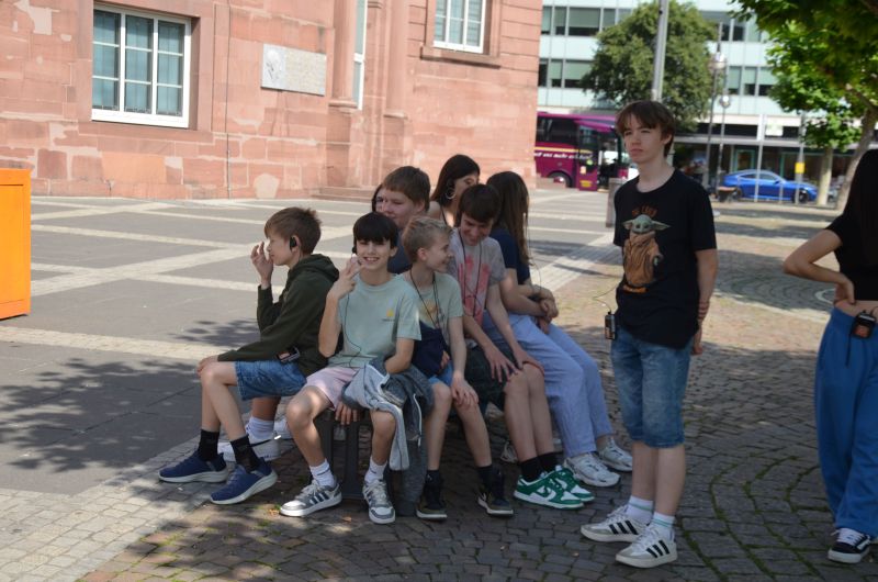 Mehrere Schüler*innen sitzen dicht gedrängt auf einer Bank vor einem alten Gebäude. Ein Junge steht daneben. Das Bild zeigt eine Pause während einer Stadtführung.