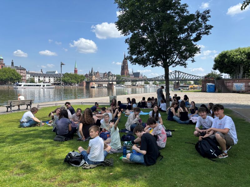Viele Jugendliche sitzen auf einer Wiese am Main. Sie picknicken und unterhalten sich.
Das Bild zeigt eine Mittagspause beim Frankfurt-Ausflug.