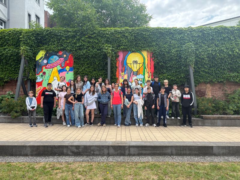 Eine große Gruppe von Schülerinnen und Schülern steht vor bunten Wandbildern mit Motiven wie einer Glühbirne und einer Rakete. Alle stehen dicht beieinander und lächeln.