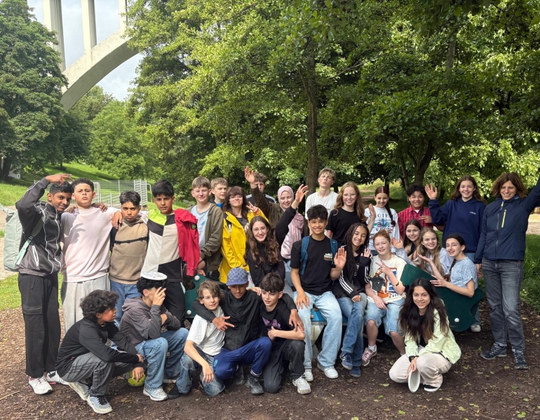Eine große Gruppe Jugendlicher steht oder sitzt im Wald unter einer Brücke. Alle winken fröhlich in die Kamera.