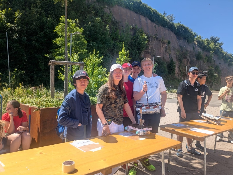 Eine Gruppe von Jugendlichen steht im Freien bei sonnigem Wetter hinter einem langen Holztisch. Sie präsentieren selbstgebaute kleine Boote, darunter auch ein Modell mit Plastikflaschen und Solarpanel.