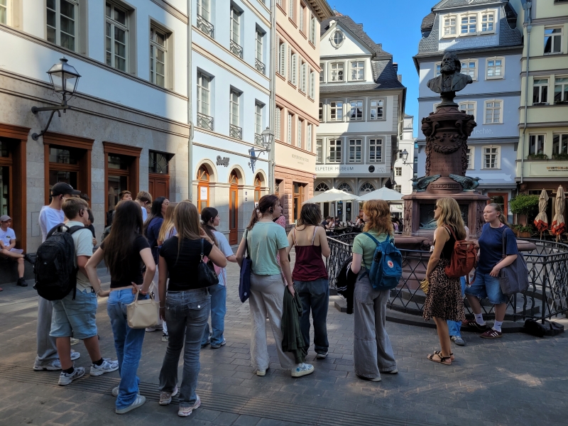 Eine Gruppe hört vor einem historischen Brunnen in der Altstadt zu. Viele tragen Rucksäcke. Das Bild zeigt eine Schulklasse bei einer Führung durch Frankfurt.