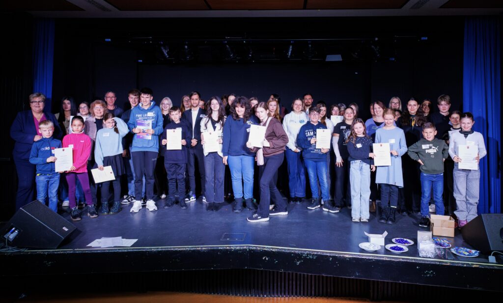 Eine große Gruppe aller Teilnehmer des Wettbewerbs stehen mit ihren Urkunden auf der Bühne.